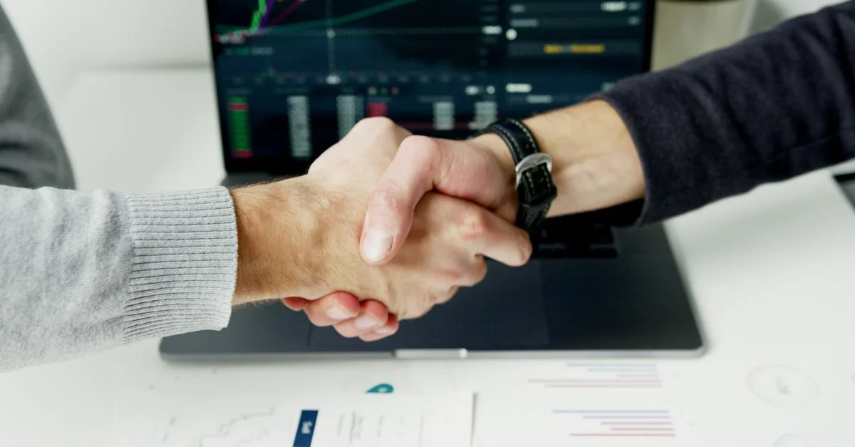Gros plan d'une poignée de main avec des graphiques financiers sur l'écran d'ordinateur portable, symbolisant un accord réussi.